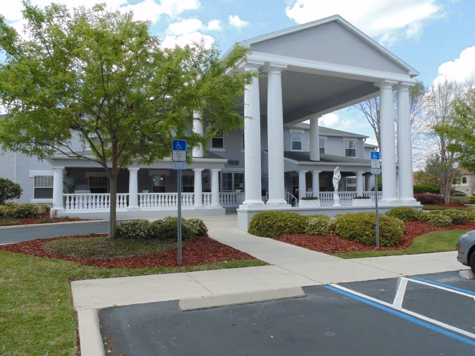Front exterior view of a senior living facility named Hampton Manor Deerwood with white columns supporting a large covered entrance, landscaped bushes and trees, and handicapped parking spaces in the foreground.