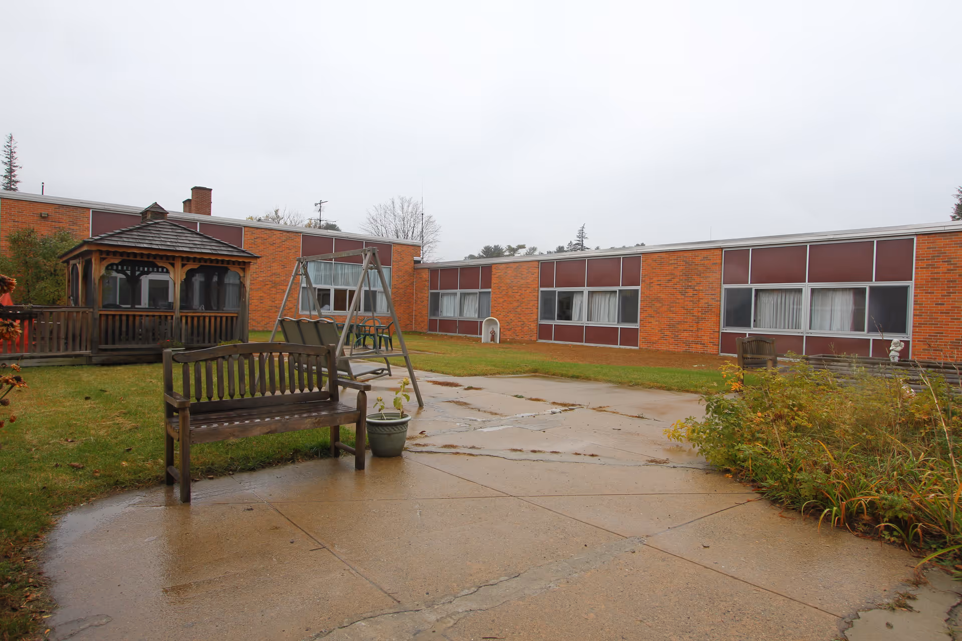 Outdoor courtyard area of Essex Center featuring a wooden gazebo, wooden benches, a swing set, and a paved walkway surrounded by grass and plants. The building with large windows and brick walls is visible in the background under an overcast sky.