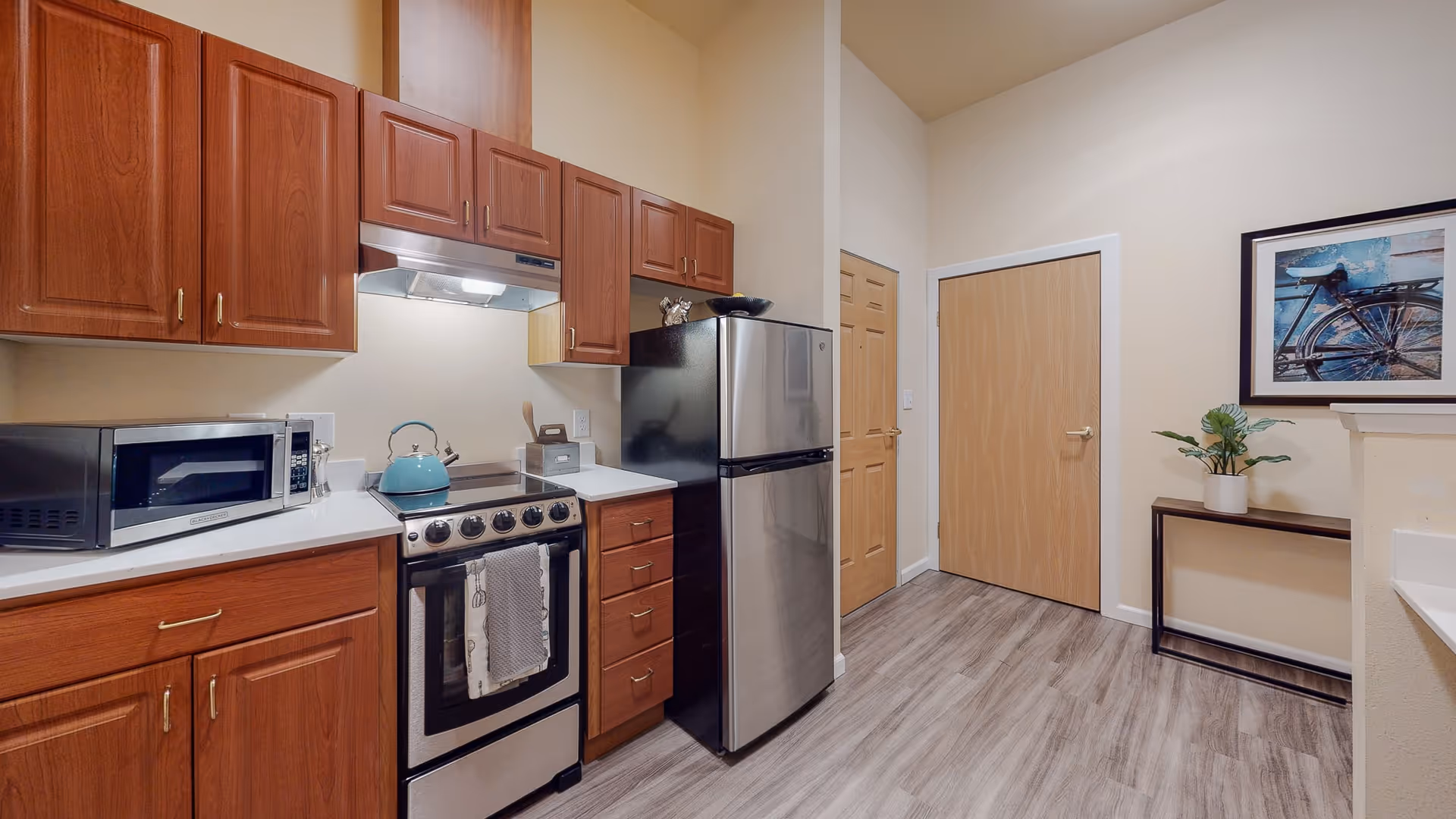 A modern kitchen with wooden cabinets, a stainless steel refrigerator, a microwave, an electric stove with a kettle on it, and a small table with a potted plant and framed bicycle artwork on the wall.