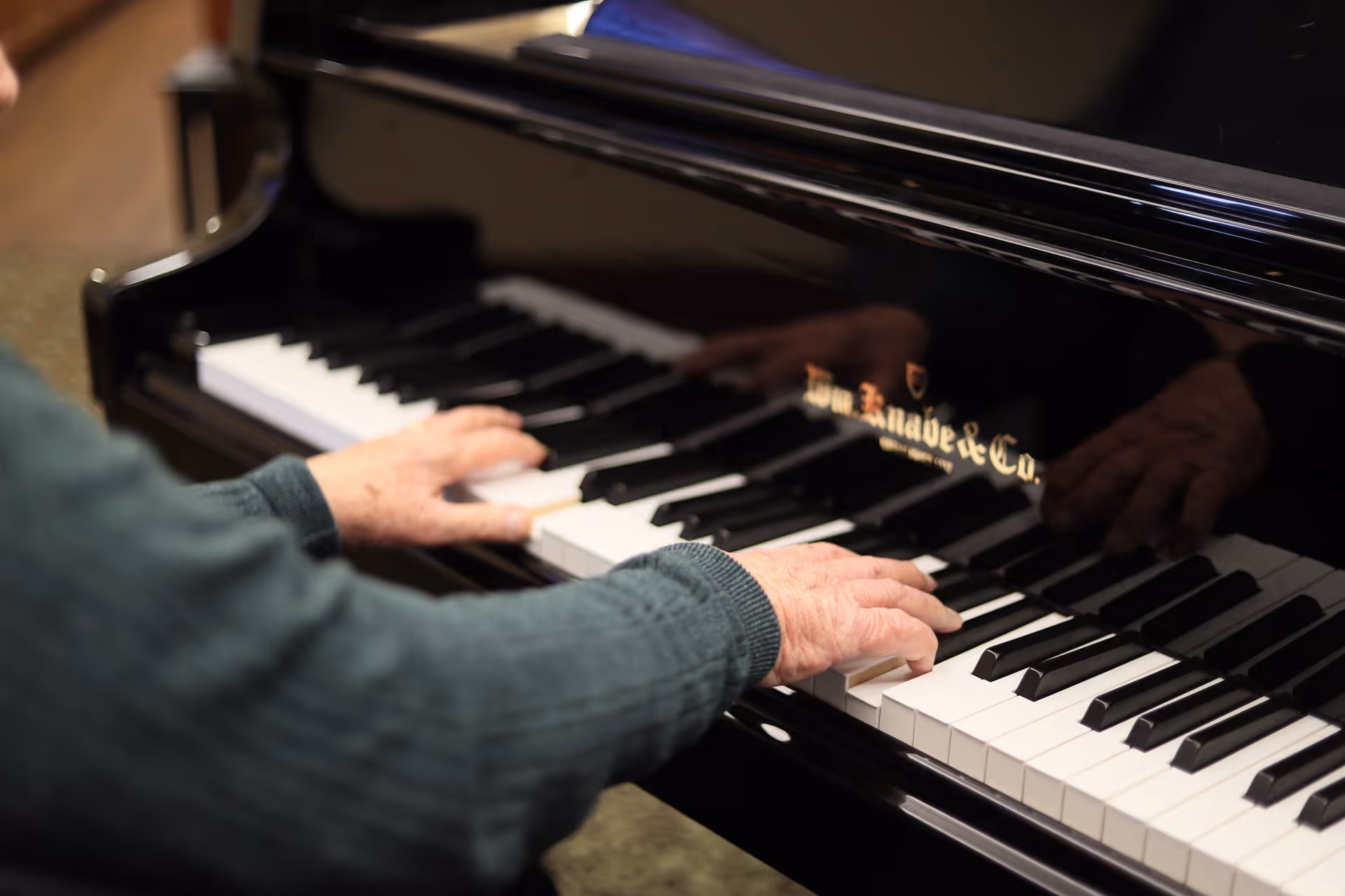 Close-up of an elderly person's hands playing a black grand piano with visible keys and the piano brand name reflected on the surface.