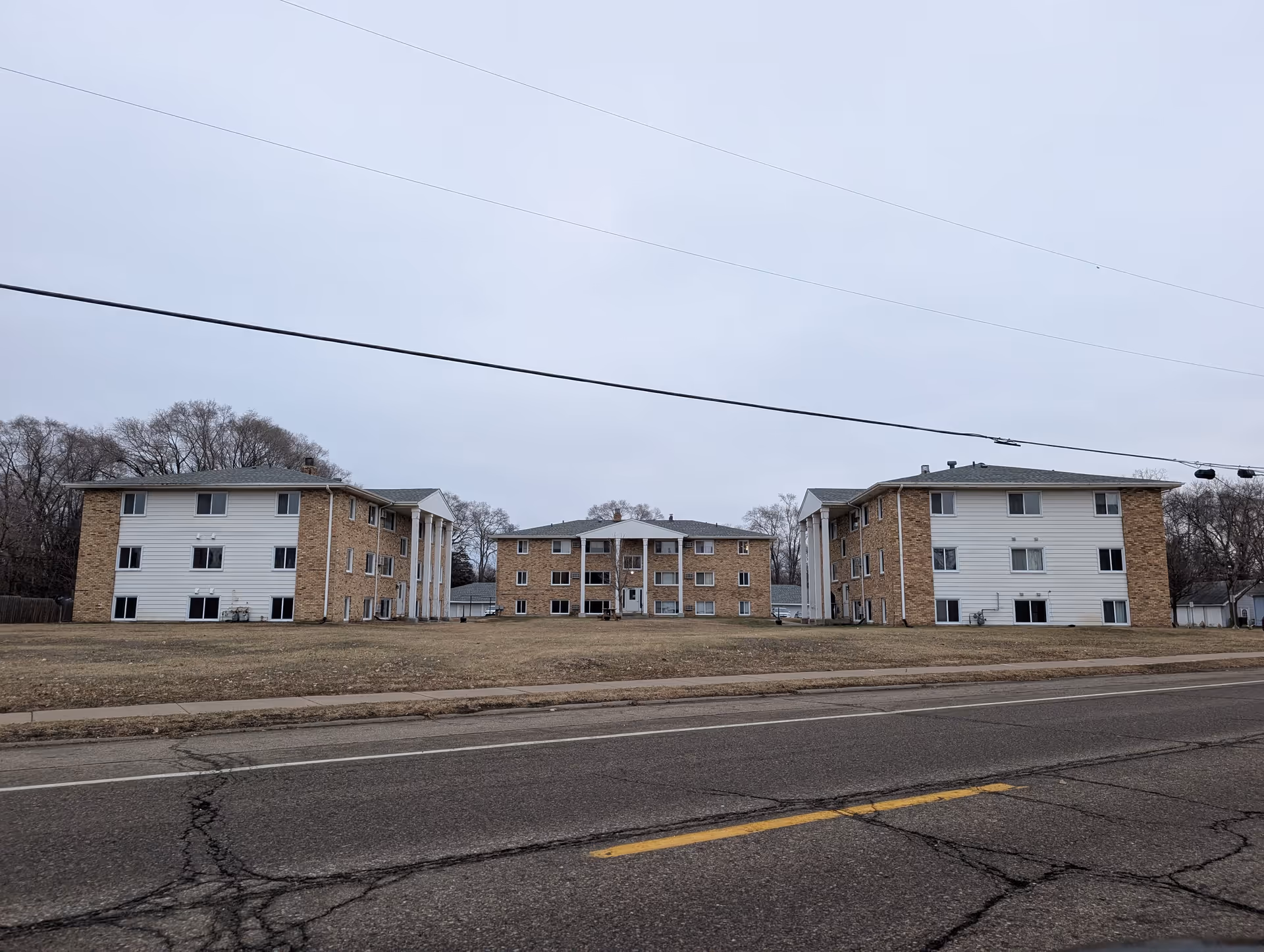 Three low-rise apartment buildings with brick and white siding exteriors, situated on a large grassy area with a cracked asphalt road in the foreground under an overcast sky.