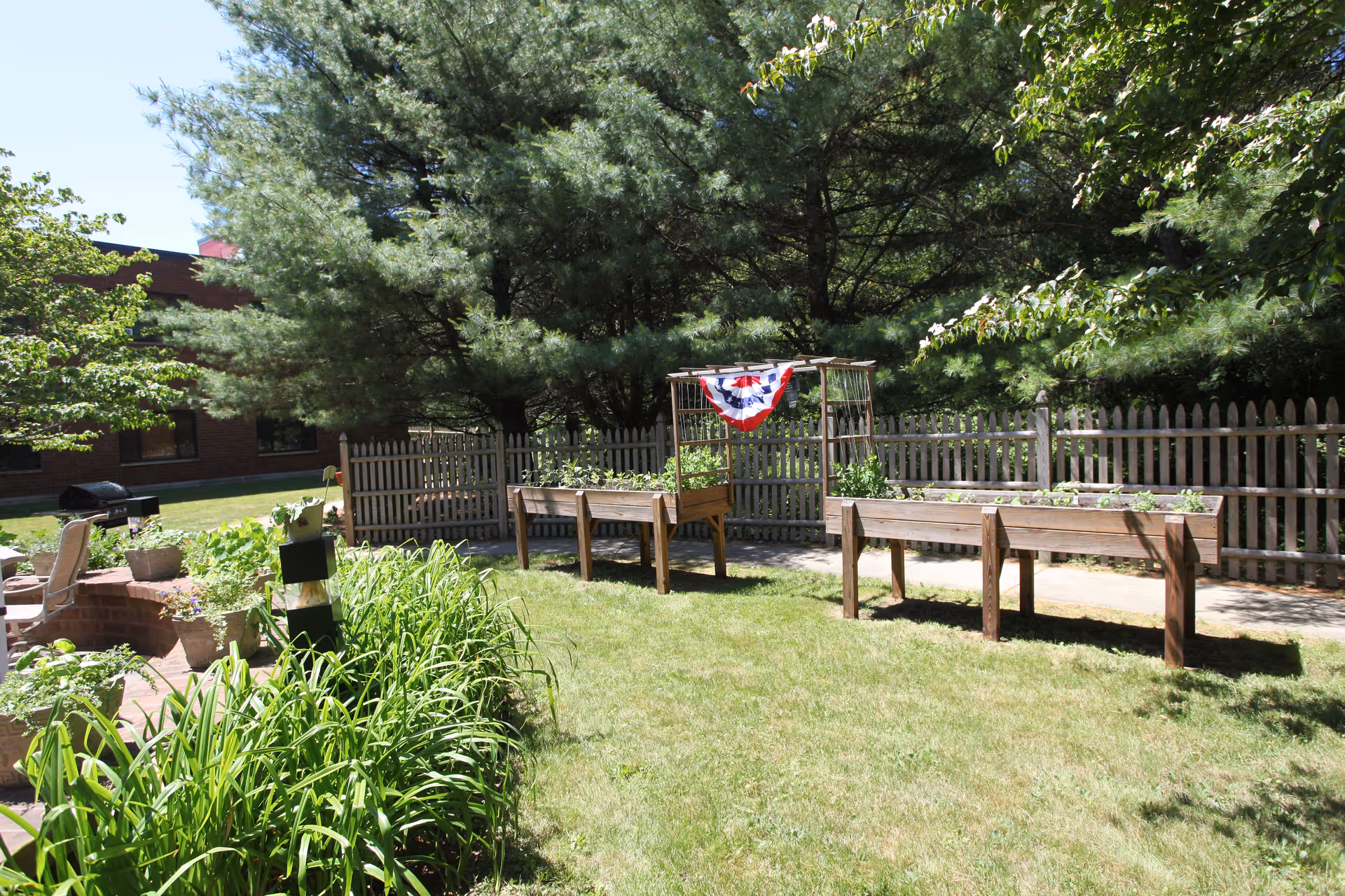 Outdoor garden area with raised wooden planter boxes containing plants, surrounded by a wooden picket fence and tall trees. There is a red, white, and blue decorative banner hanging on a wooden trellis. The area is grassy with some potted plants and a brick patio with chairs visible on the left side.