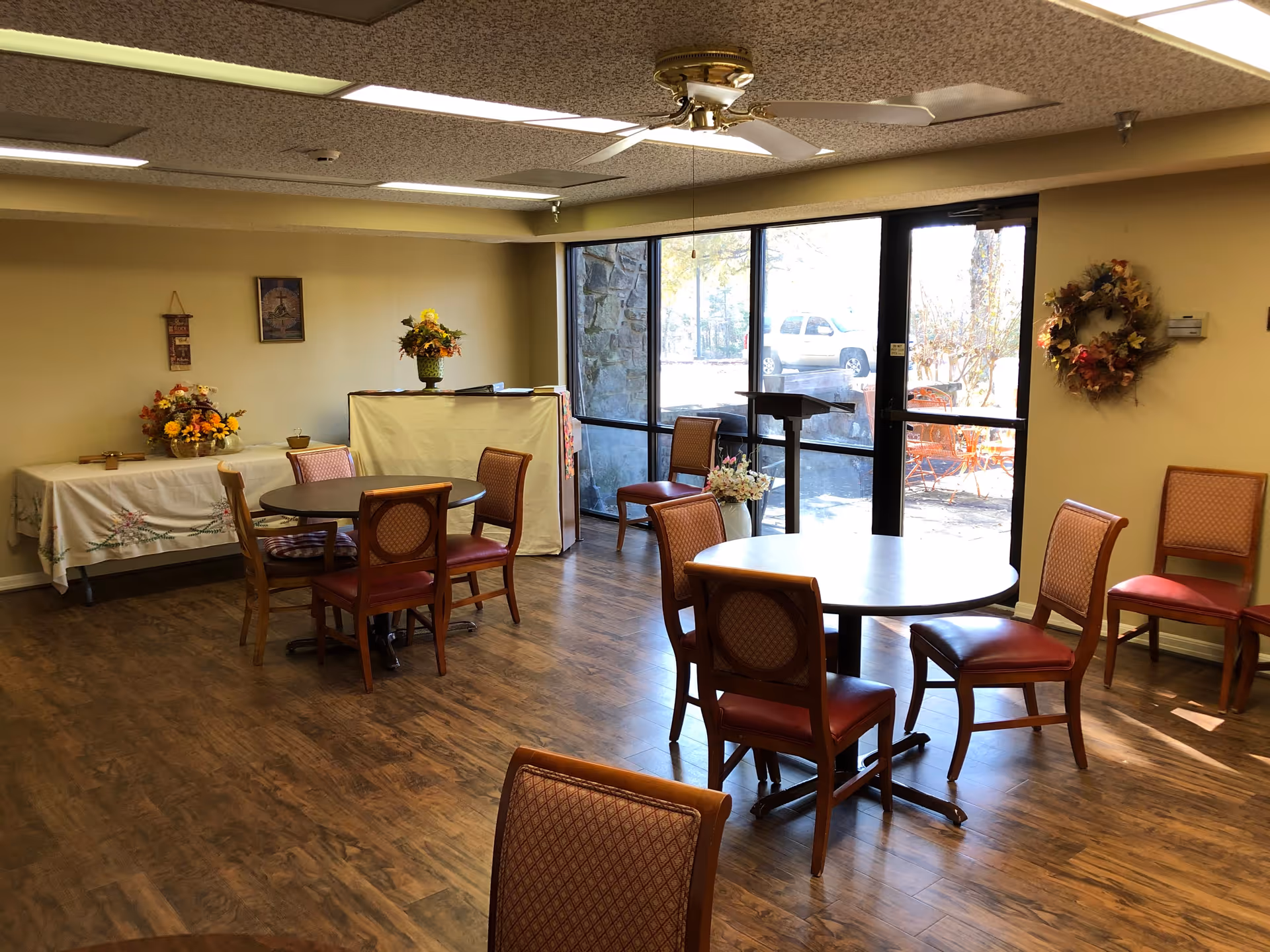 A well-lit room with wooden flooring featuring two round tables surrounded by chairs with red cushioned seats. There is a large window and glass door letting in natural light, with a view of outdoor seating and a parked vehicle. The walls are decorated with floral wreaths and framed pictures, and there are flower arrangements on a covered table and a stand.