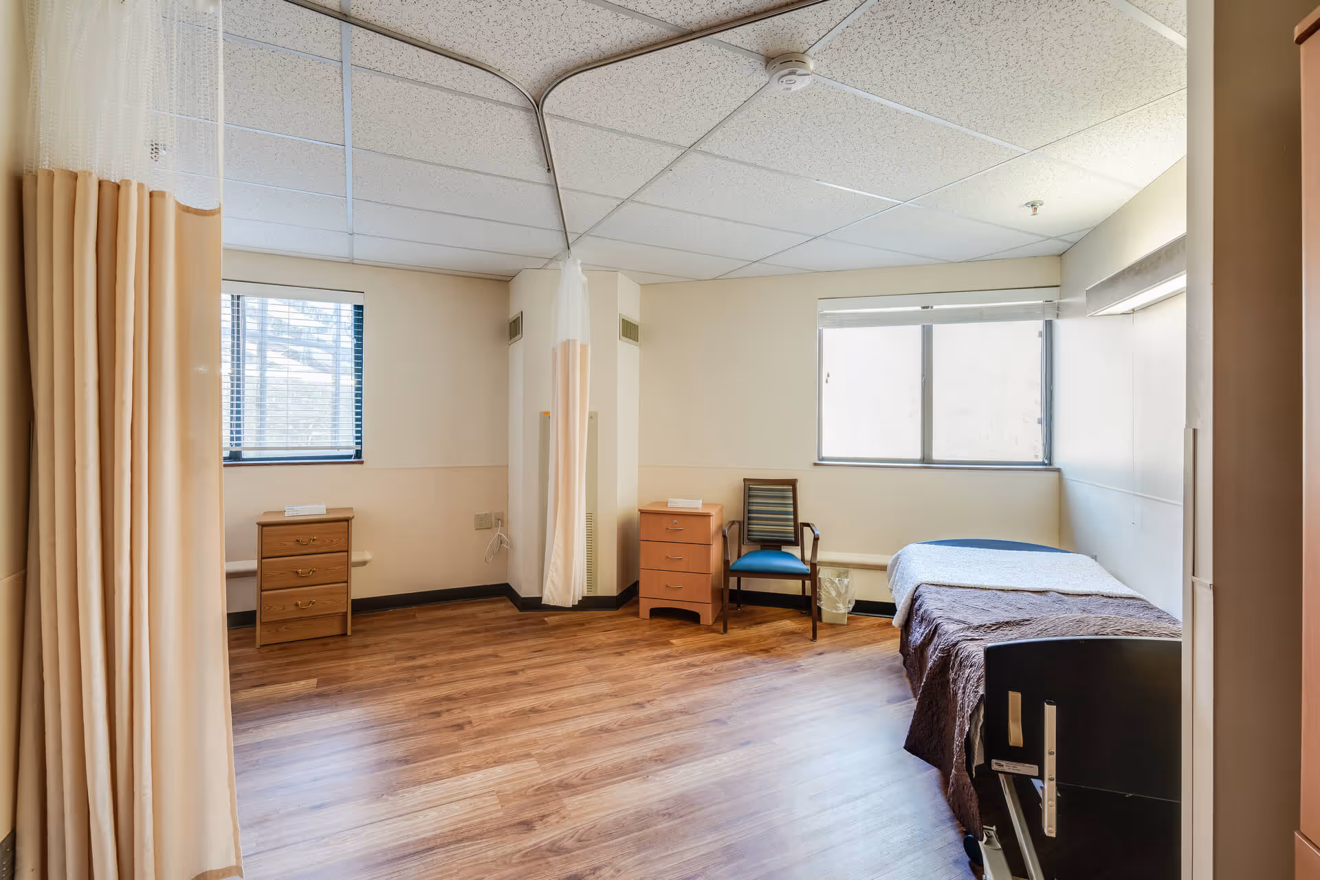 Simple senior living bedroom with a single bed, bedside tables, a chair, privacy curtains, and wood-look flooring.