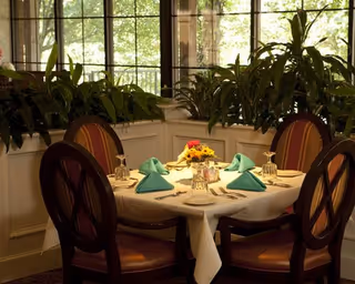 A round dining table set for six with a white tablecloth, teal folded napkins, glassware, and a centerpiece of yellow and red flowers. The table is surrounded by wooden chairs with cushioned seats and backs. Large windows and green plants are visible in the background.
