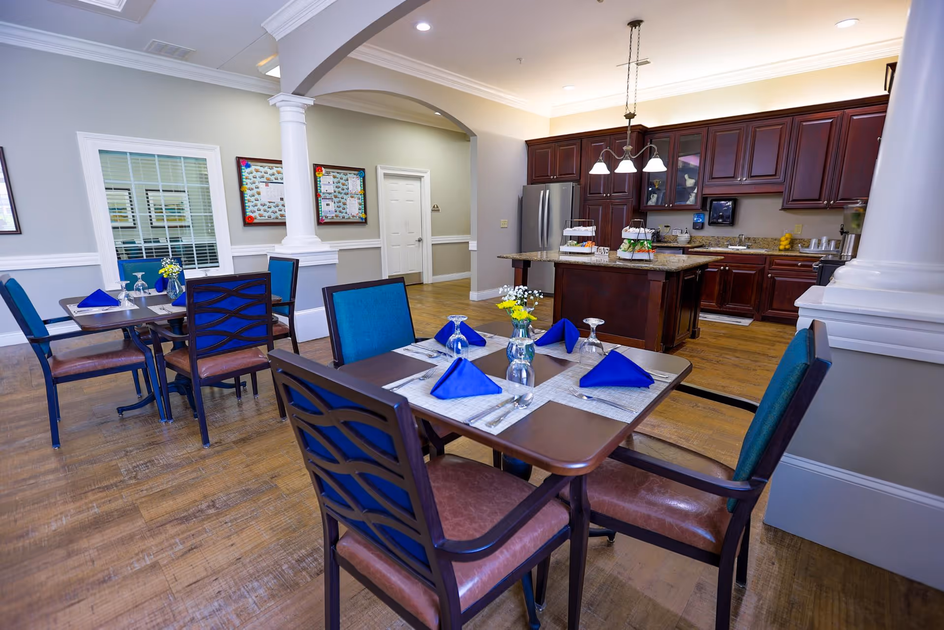 Dining area with tables set with blue napkins, glassware, and small flower vases. The room features wooden floors, a kitchen area with dark wood cabinets, a refrigerator, and an island with food trays. The walls are light-colored with white trim and columns.
