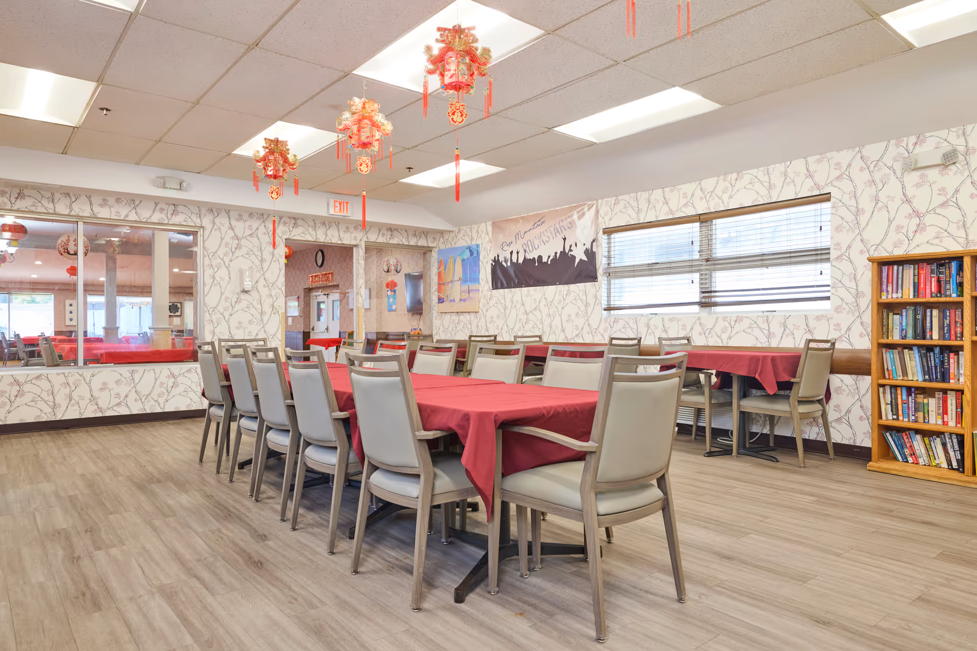 A dining room with several tables covered in red tablecloths and surrounded by beige chairs. The room has light wood flooring, floral patterned wallpaper, and fluorescent ceiling lights. Red hanging decorations are suspended from the ceiling. There is a bookshelf filled with books on the right side and a window with blinds. A banner and some artwork are visible on the walls.