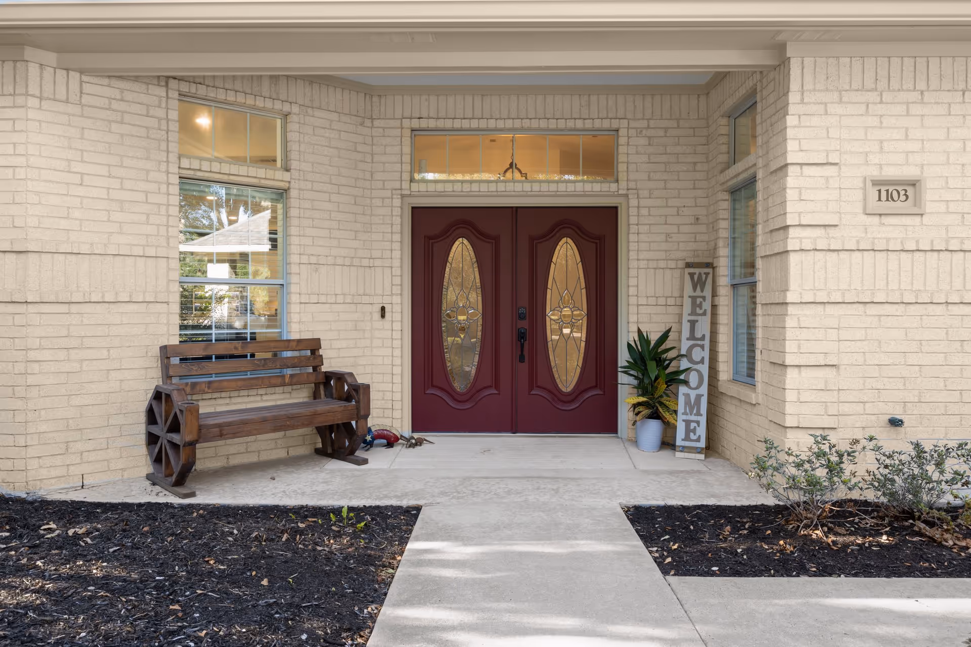 Front entrance with double red decorative doors, a 'WELCOME' sign, wooden bench, and address plaque on a brick facade.