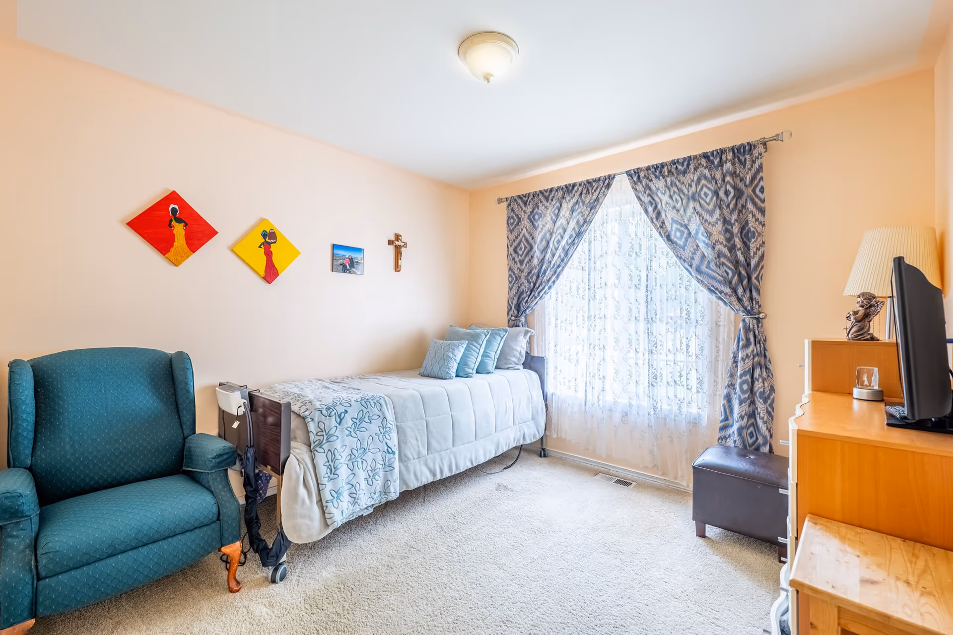 A cozy senior care bedroom with a single bed covered in a light blue and white comforter and several pillows. Next to the bed is a teal upholstered armchair. The room has beige walls adorned with colorful artwork and a small cross. A window with sheer white curtains and patterned blue drapes lets in natural light. A wooden dresser with a TV and a lamp is positioned opposite the bed.