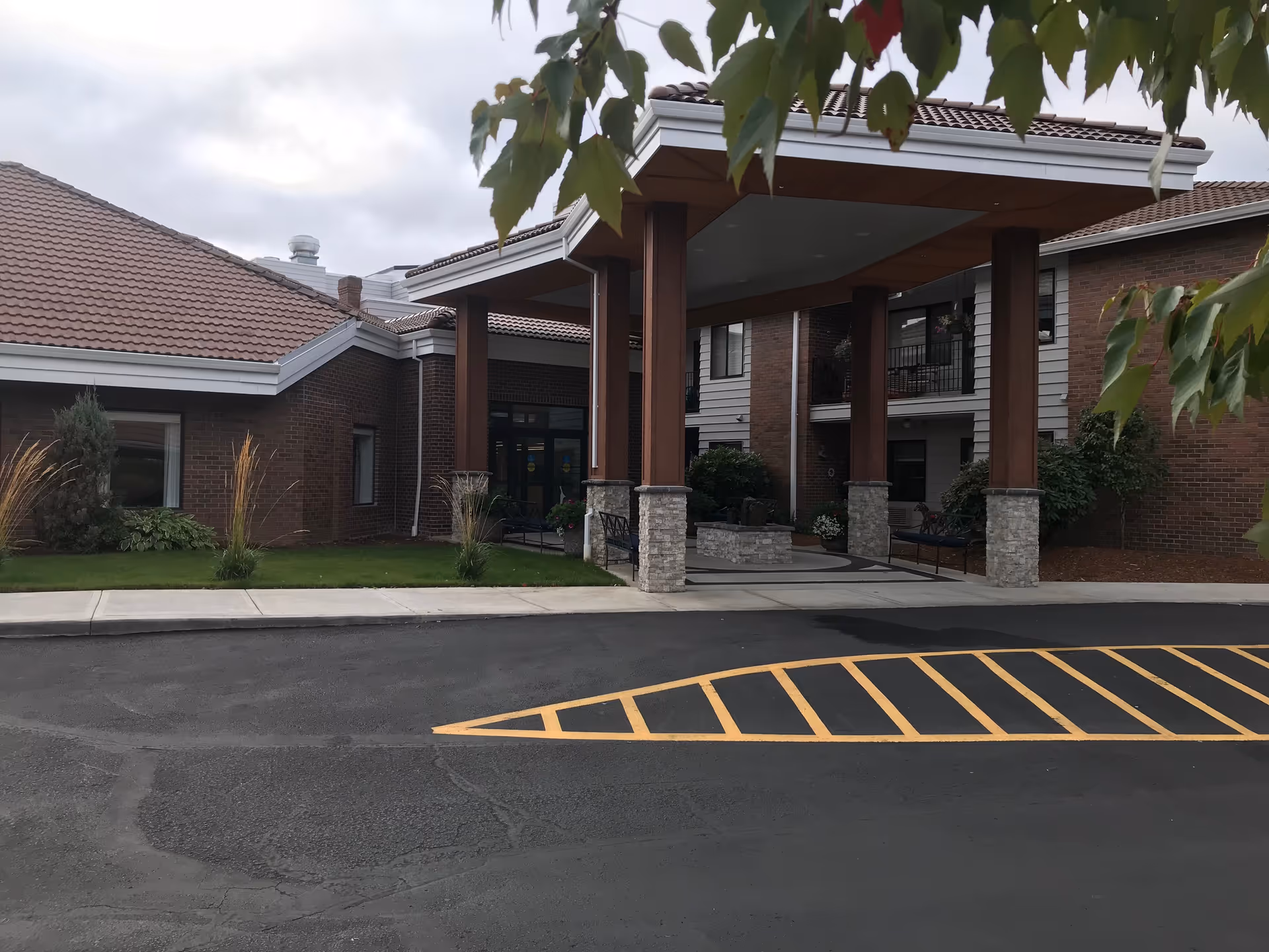 Entrance area of a senior living facility with a covered drop-off zone supported by wooden pillars with stone bases. The building has brick walls and a tiled roof, with some greenery and trees partially visible in the foreground.