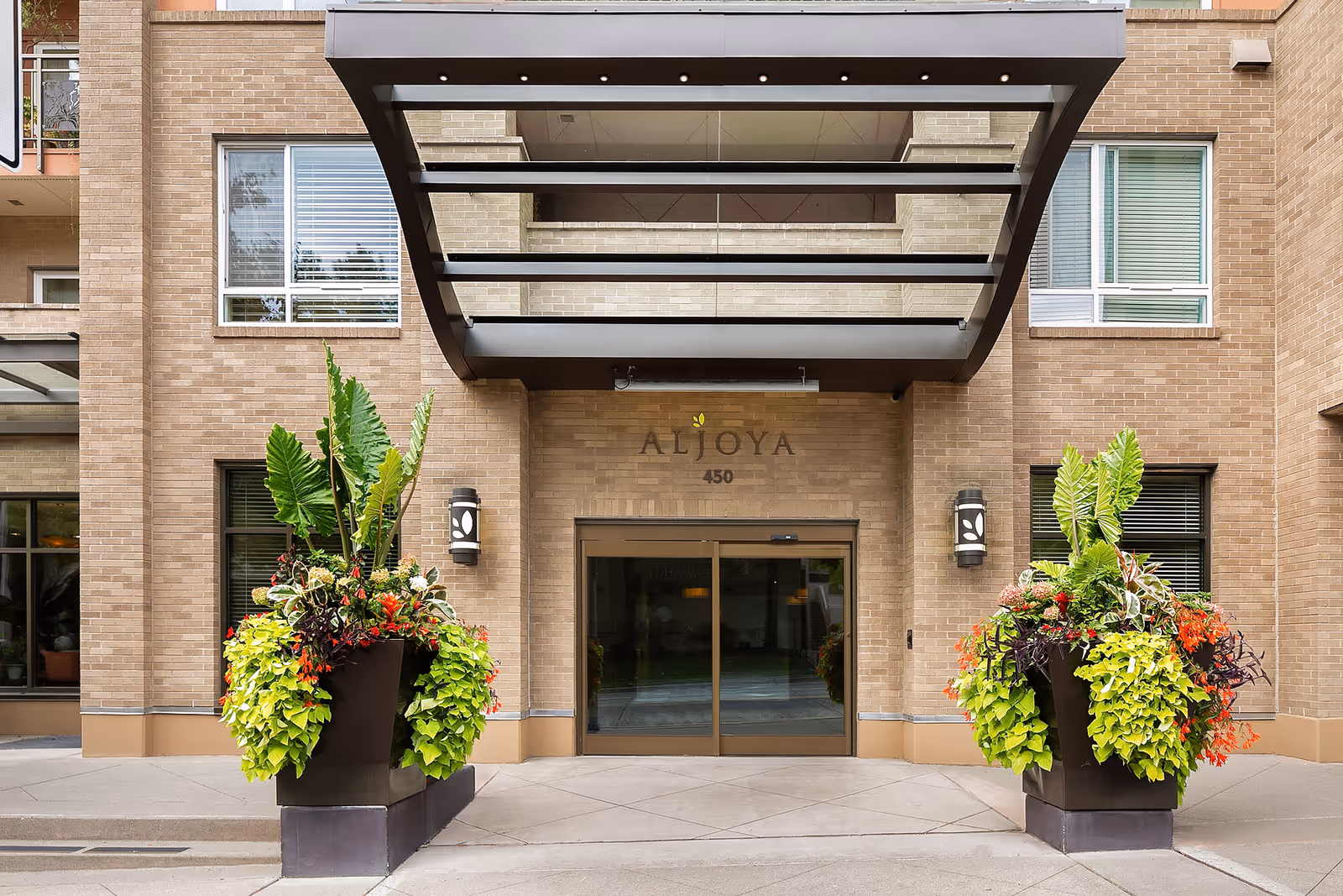 Entrance to Aljoya Thornton Place facility with a glass door under a modern metal and glass canopy. Two large planters with green and red foliage flank the entrance. The building exterior is made of light brown brick with windows on either side of the door.