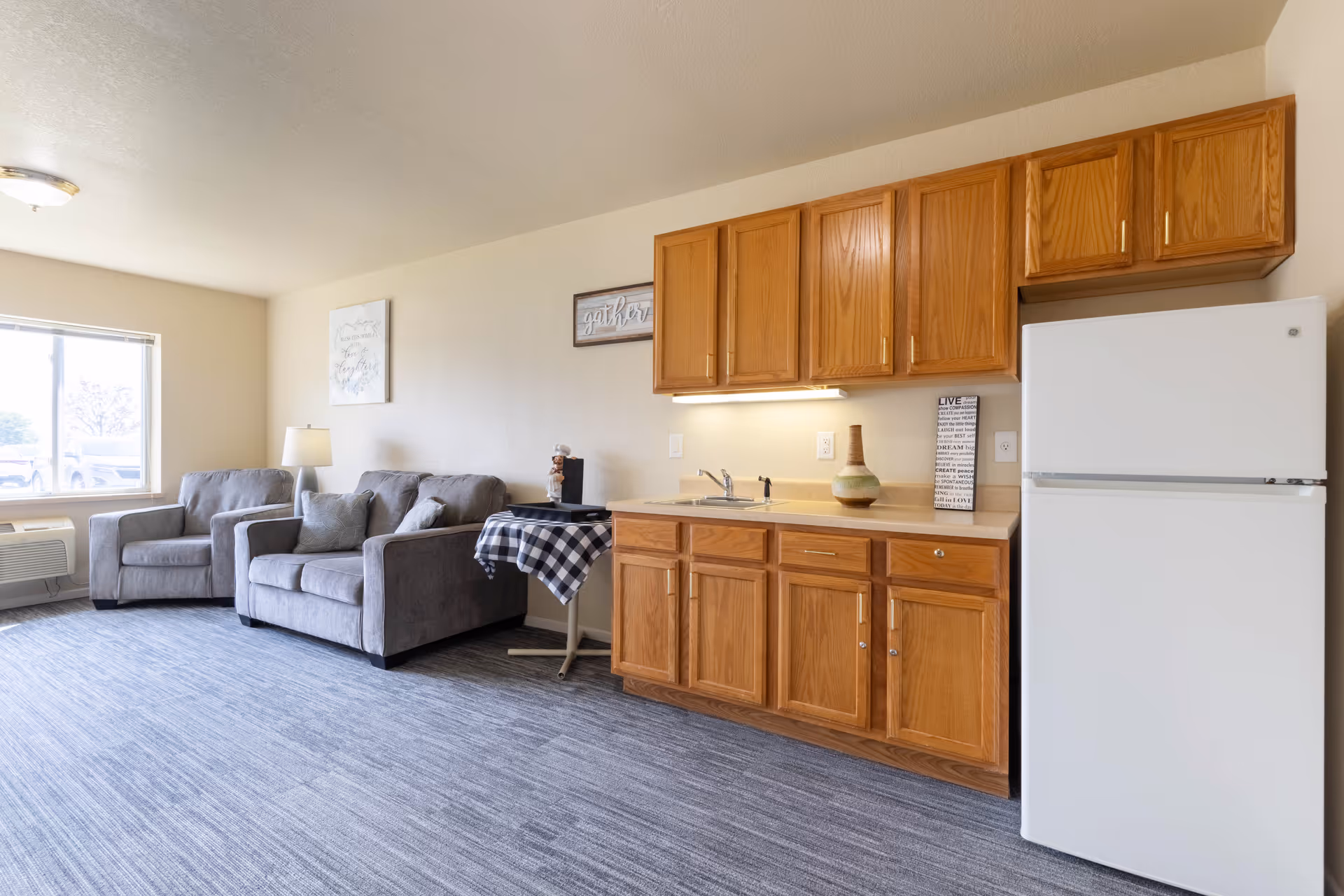 A bright room featuring a small kitchen area with wooden cabinets, a white refrigerator, and a countertop with a sink. Adjacent to the kitchen is a living area with a gray loveseat and armchair, a small table covered with a black and white checkered cloth, a lamp, and wall art. A large window allows natural light to fill the space.