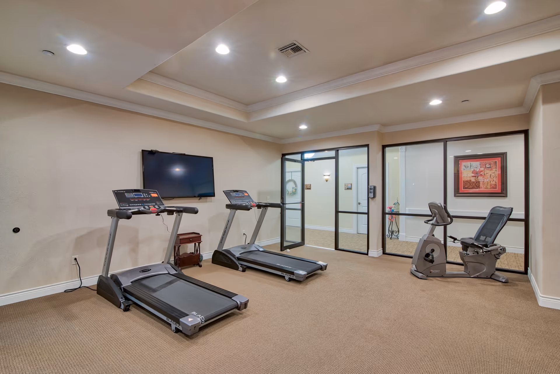 A fitness room with two treadmills and a recumbent exercise bike. The room has beige walls, carpeted floor, recessed ceiling lights, a wall-mounted TV, and glass doors leading to another area with framed artwork on the walls.