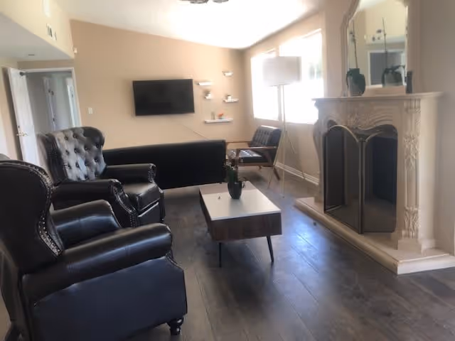 A living room with two black leather armchairs, a black sofa, a wooden coffee table with a small plant, a fireplace with a decorative mantel and mirror above it, a floor lamp, and a window letting in natural light.