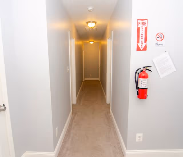A narrow indoor hallway with beige carpet and light gray walls, featuring several doorways on both sides. The ceiling has two light fixtures, and a red fire extinguisher is mounted on the right wall below a fire extinguisher sign and a no smoking sign.
