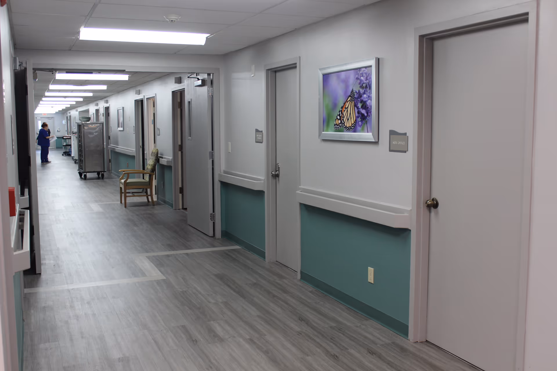 A clean, well-lit hallway in a senior care facility with light gray walls and teal lower wall panels. Several closed doors line the hallway, one labeled 'Nurse Office'. A framed picture of a butterfly on purple flowers hangs on the wall. A chair is placed along the hallway and a staff member in blue scrubs is seen in the distance near a metal cart.