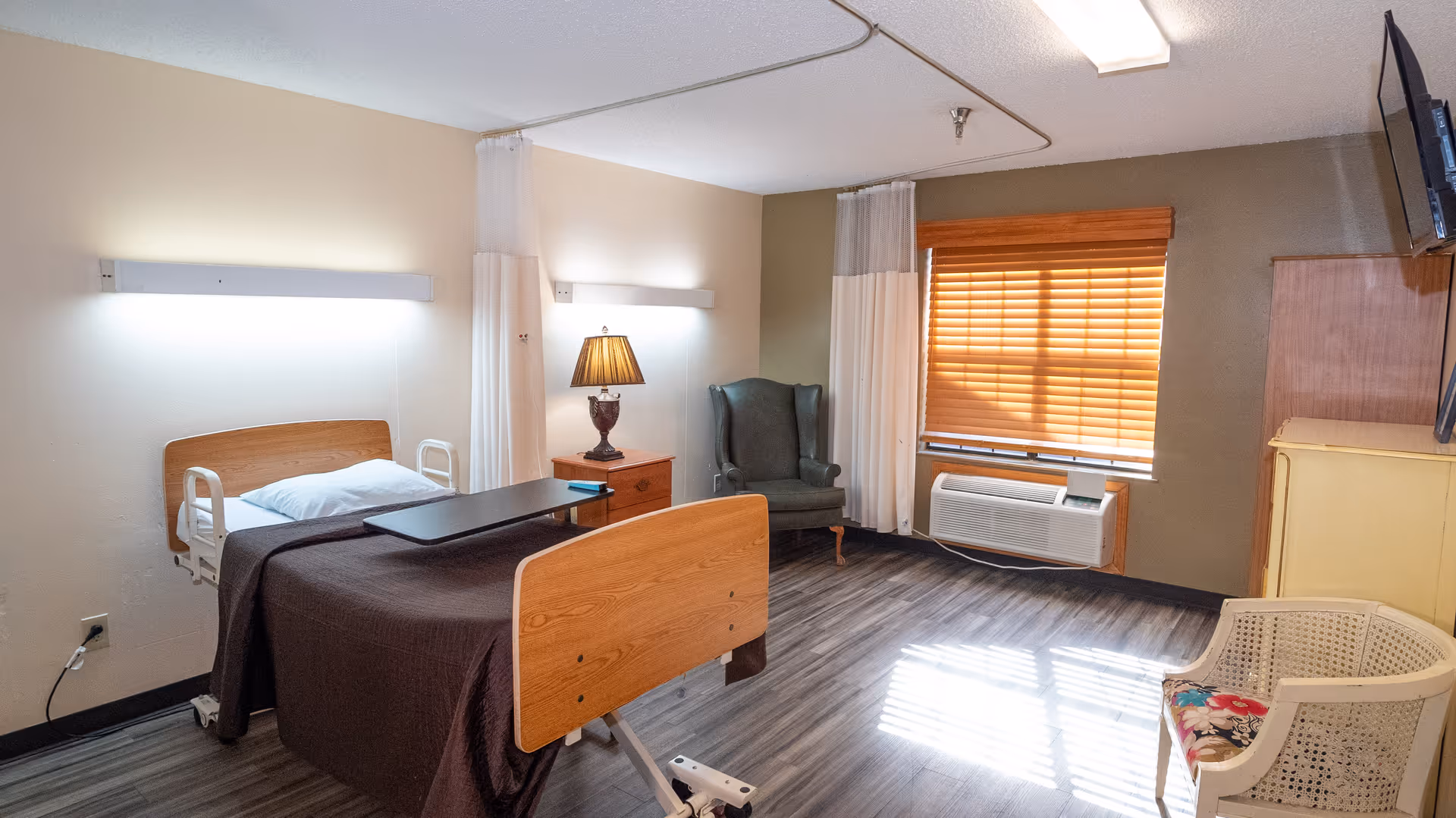 A senior care patient room with a hospital bed, overbed table, armchair, lamp, and a window with blinds.