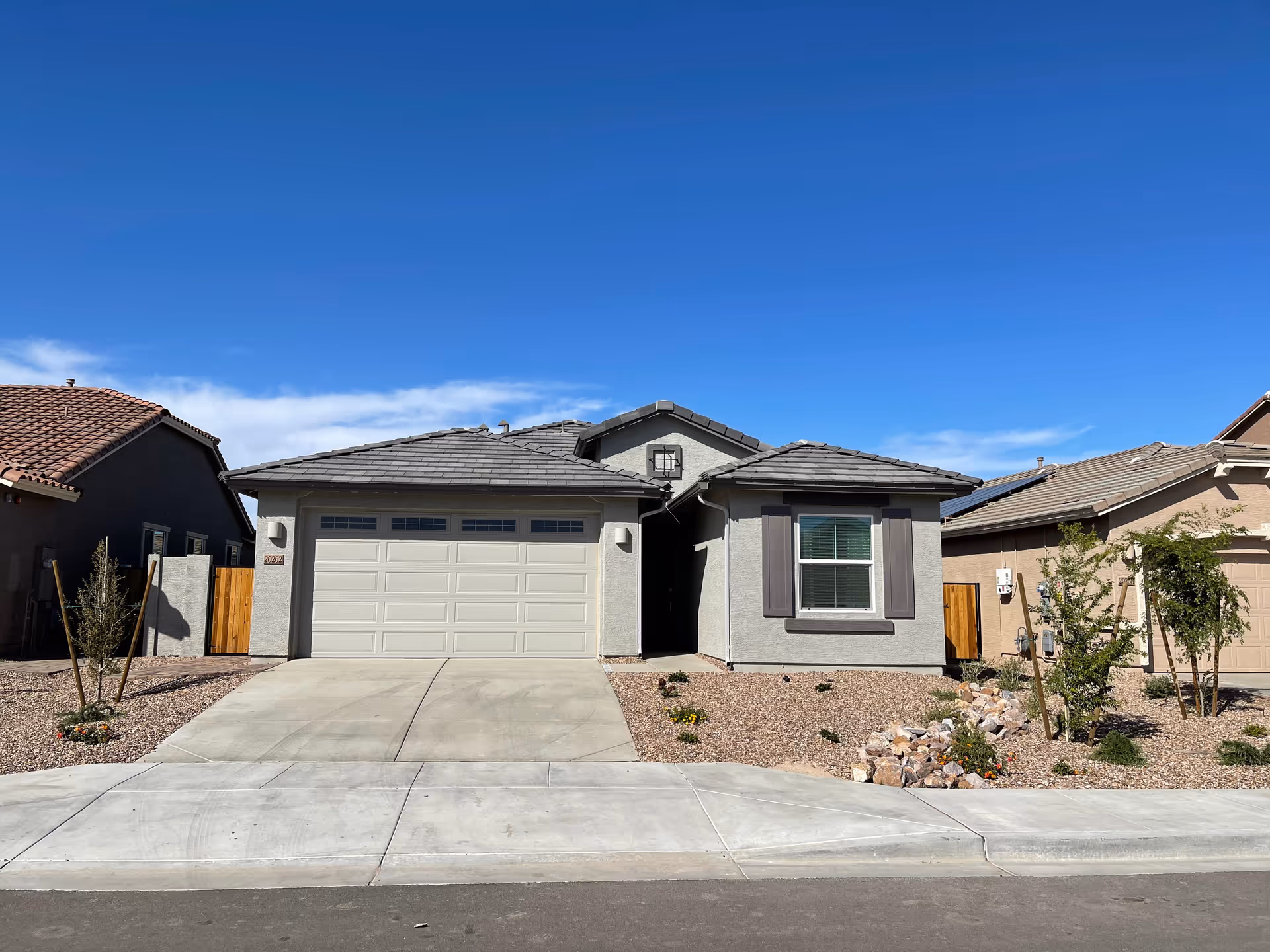 Front exterior view of a single-story house with a two-car garage, light gray walls, dark gray roof tiles, and a small landscaped front yard with rocks and plants under a clear blue sky.