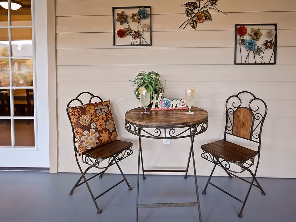 Bistro table with two decorative metal chairs, drinks, a 'Home' sign and floral wall decor against a paneled exterior wall.