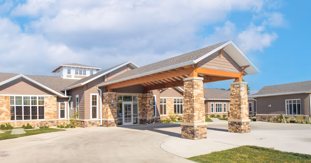 Front exterior view of Grand Meadows Senior Living facility showing a covered entrance with stone pillars, large windows, and a clear blue sky with some clouds.