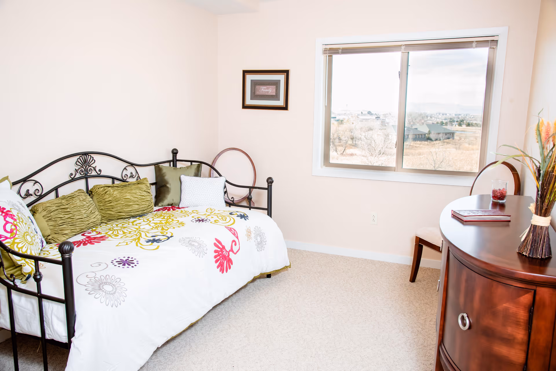 Bright bedroom with a decorative metal daybed covered in floral bedding, throw pillows, a window, and a wooden dresser.