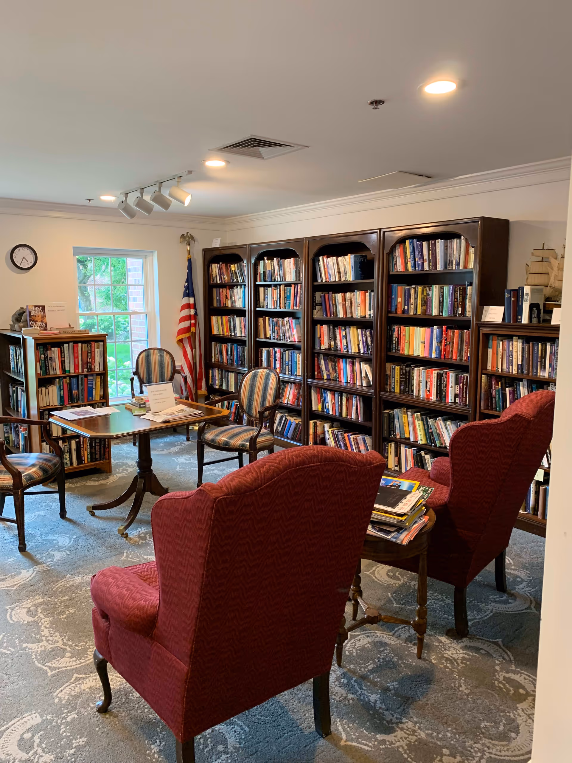 A cozy library room with multiple wooden bookshelves filled with books along the walls. There are two red upholstered armchairs facing a small wooden table with magazines on it. In front of the bookshelves, there is a wooden table surrounded by four striped upholstered chairs. A window lets in natural light, and an American flag stands in the corner. The room has a patterned carpet and ceiling lights.