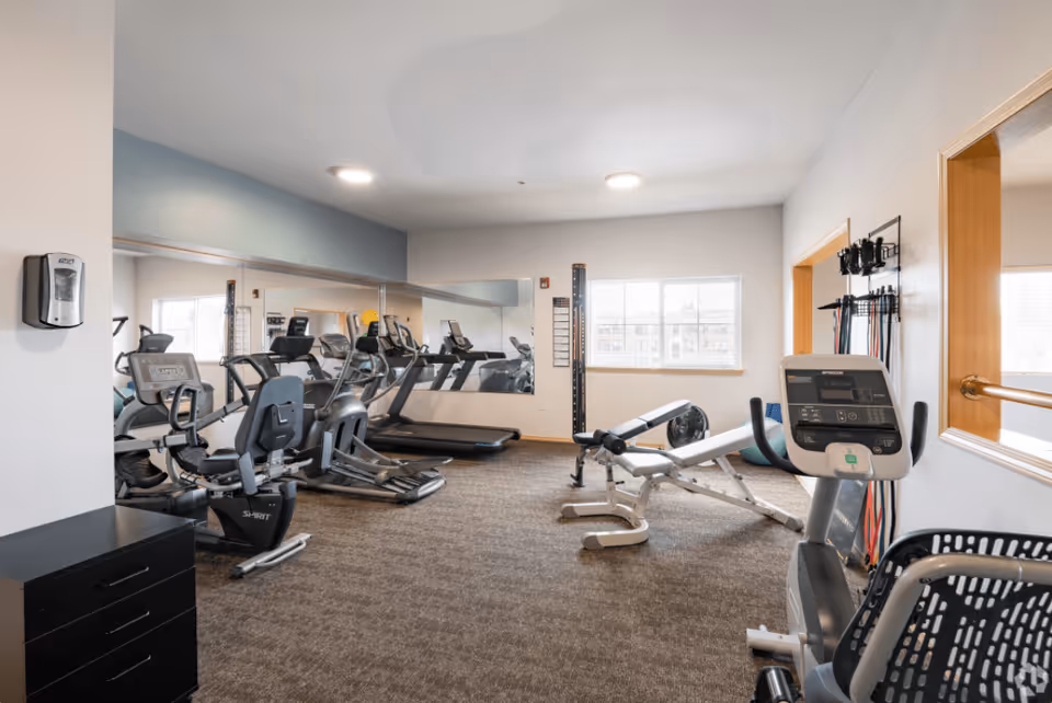 A fitness room with exercise equipment including treadmills, stationary bikes, a weight bench, and resistance bands mounted on the wall. The room has carpeted flooring, a large mirror on one wall, and a window letting in natural light.