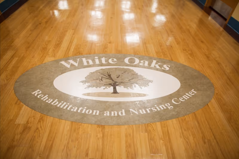 A polished wooden floor with an oval-shaped floor mat in the center displaying the logo and name 'White Oaks Rehabilitation and Nursing Center' with an image of a tree.