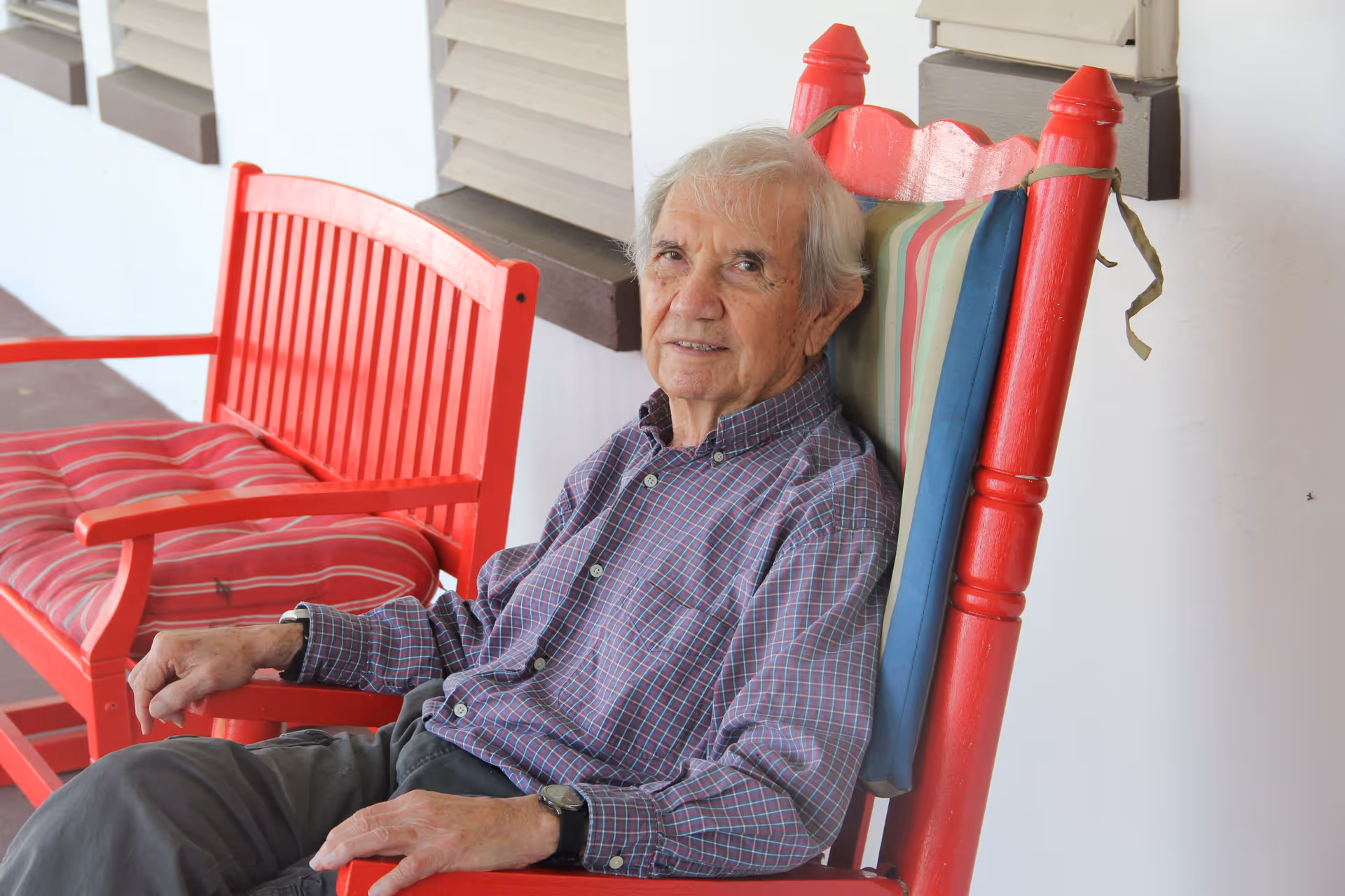 An elderly man with gray hair wearing a checkered shirt and dark pants is sitting on a red wooden rocking chair with a striped cushion on the backrest. Next to him is a matching red wooden bench with a red striped cushion. They are on a porch with white walls and windows with brown shutters.