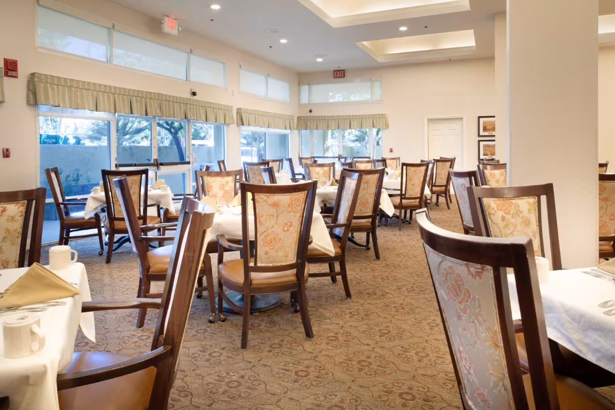 Well-lit dining room with multiple tables and upholstered wooden chairs set with tablecloths and place settings.