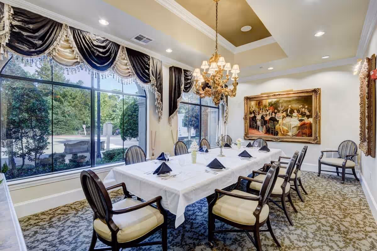 Elegant dining room with a long table covered with a white tablecloth, set with black napkins and glassware. The room features large windows with decorative drapes, a chandelier hanging from a tray ceiling, patterned carpet, and framed artwork on the walls.