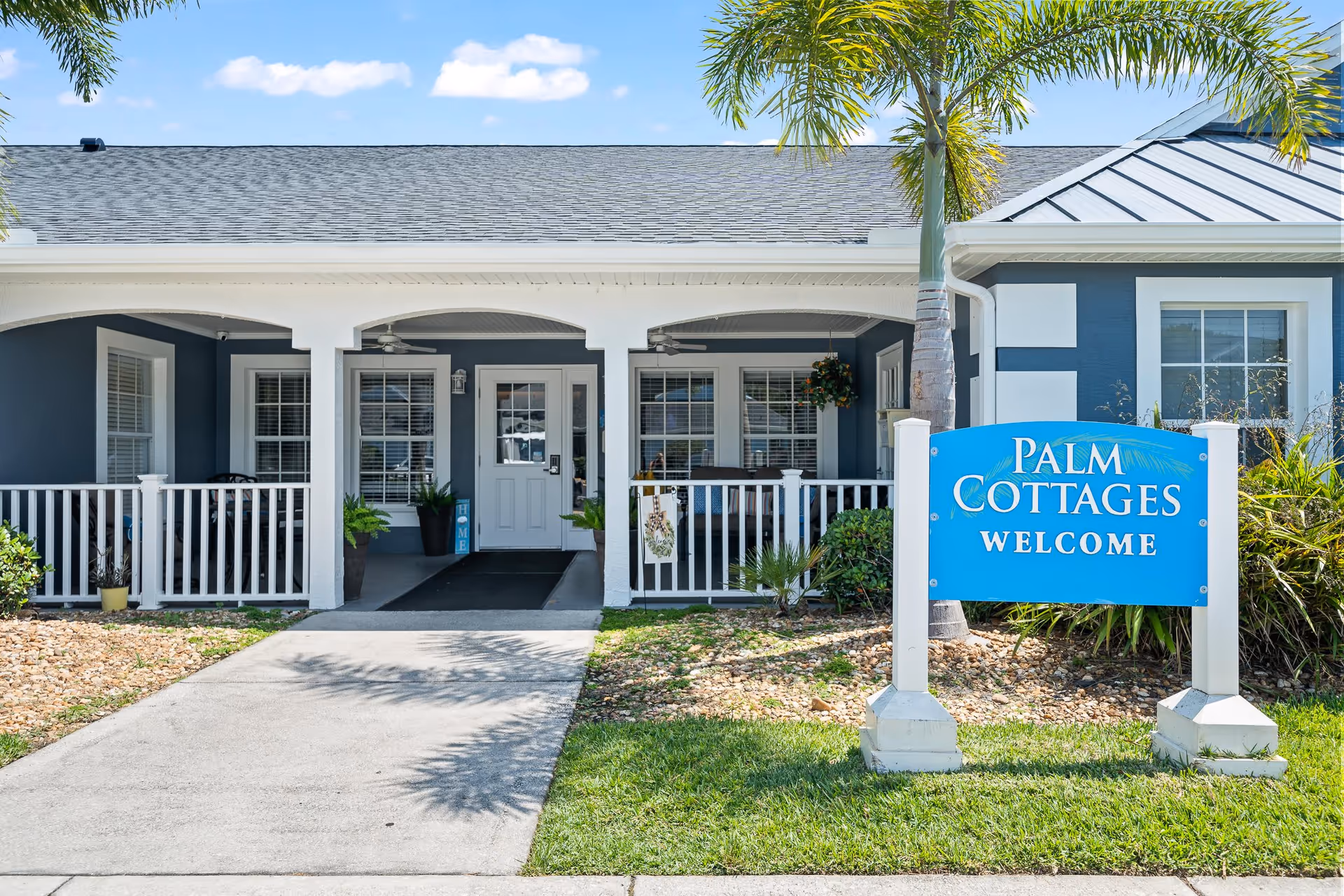 Front exterior view of Palm Cottages of Rockledge showing a covered porch with white railings, potted plants, and a blue welcome sign on the lawn. The building has a gray and white color scheme with a shingled roof and palm trees nearby.