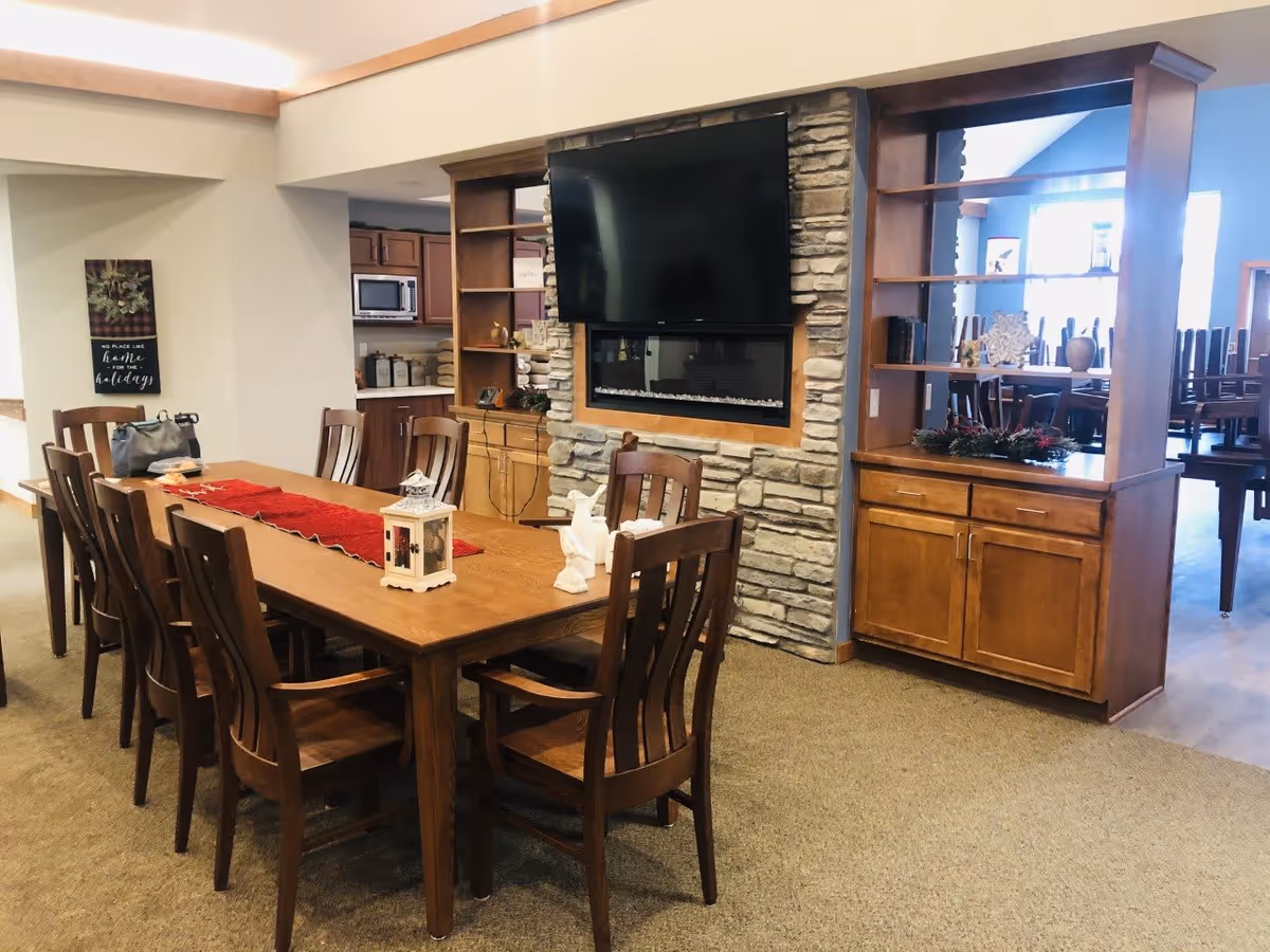 A cozy dining area with a long wooden table surrounded by wooden chairs. The table is decorated with a red runner and small decorative items including a white lantern and a white bird figurine. Behind the table is a stone wall with a mounted flat-screen TV above an electric fireplace. To the right, there is a wooden shelving unit with cabinets and decorative items. In the background, a kitchen area with wooden cabinets and a microwave is visible.