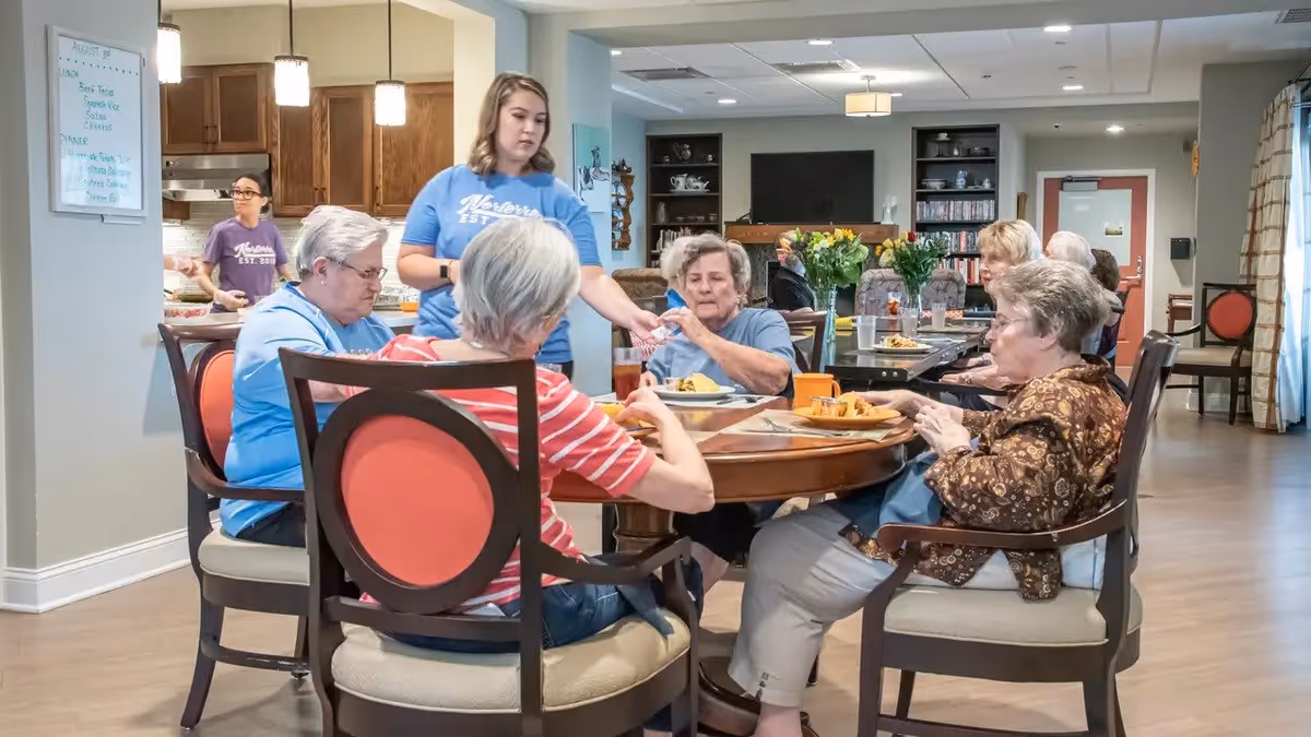 A group of elderly women sitting around a round wooden table in a dining area, eating a meal. A caregiver in a blue shirt is serving or assisting them. The room has wooden flooring, a kitchen area in the background, and a cozy living space with a TV and shelves.