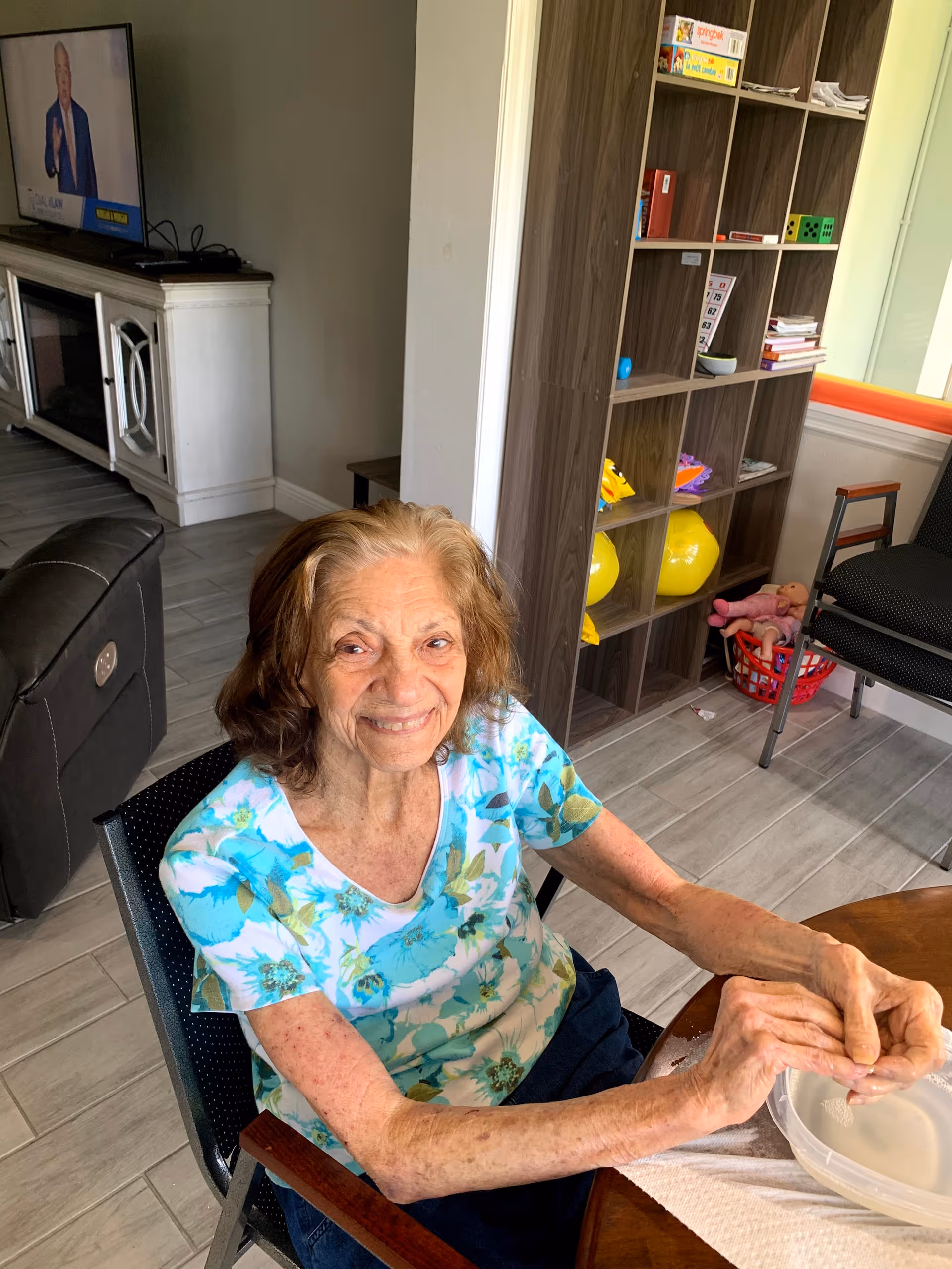 An elderly woman with light brown hair wearing a floral blue and white shirt is sitting at a round wooden table in a room. She is smiling and holding a plastic container. Behind her, there is a shelving unit with various items including yellow balloons and board games. A television is on in the background, and there are chairs and a couch visible in the room.
