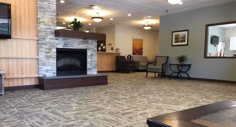 A spacious and well-lit common area with a modern stone fireplace, patterned carpet, and comfortable seating including armchairs and a small table with a plant. The room has neutral-colored walls with framed artwork and a large mirror reflecting part of the room. Ceiling lights provide warm illumination.