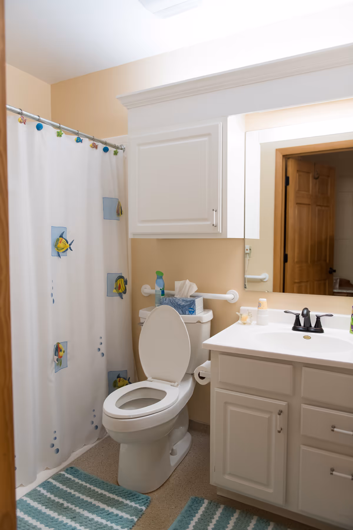 A clean bathroom with a white toilet, a white vanity with a sink and black faucet, a large mirror, and a white cabinet above the toilet. The shower has a white curtain decorated with colorful fish and sea creatures. There are two blue and white striped bath mats on the floor.