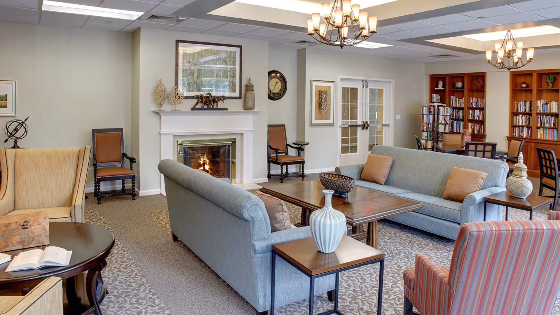 A cozy living room area in a senior living facility featuring two blue sofas with beige pillows, a striped armchair, a beige armchair, a wooden coffee table, side tables with decorative vases, a lit fireplace, framed artwork on the walls, and bookshelves filled with books in the background.