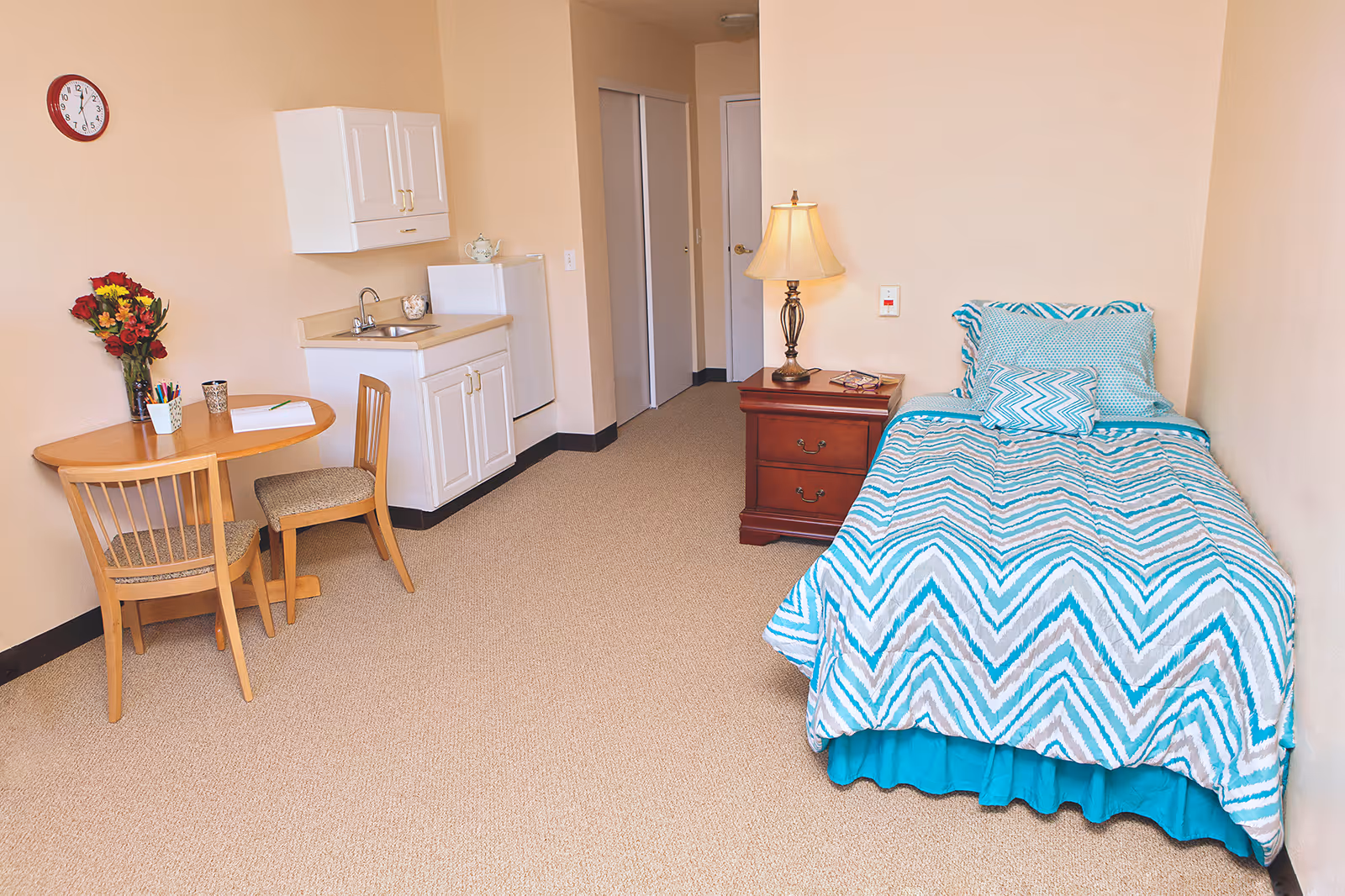 Small studio room with a single bed in teal chevron bedding, a nightstand and lamp, a kitchenette and a round table with two chairs.