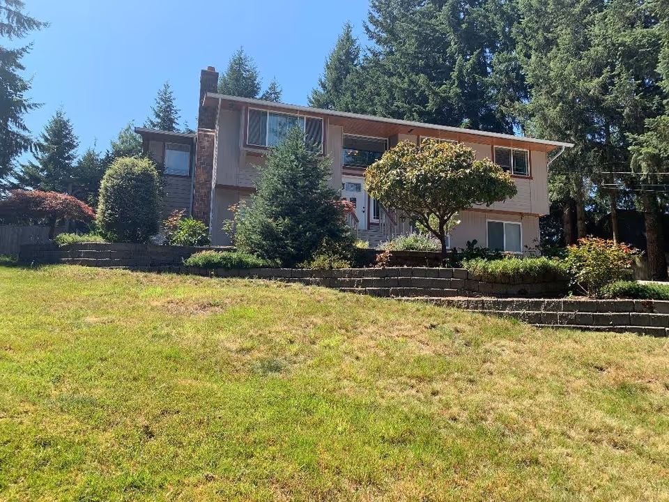 Two-story house front with terraced landscaping, trees, and a grassy lawn under a clear sky.