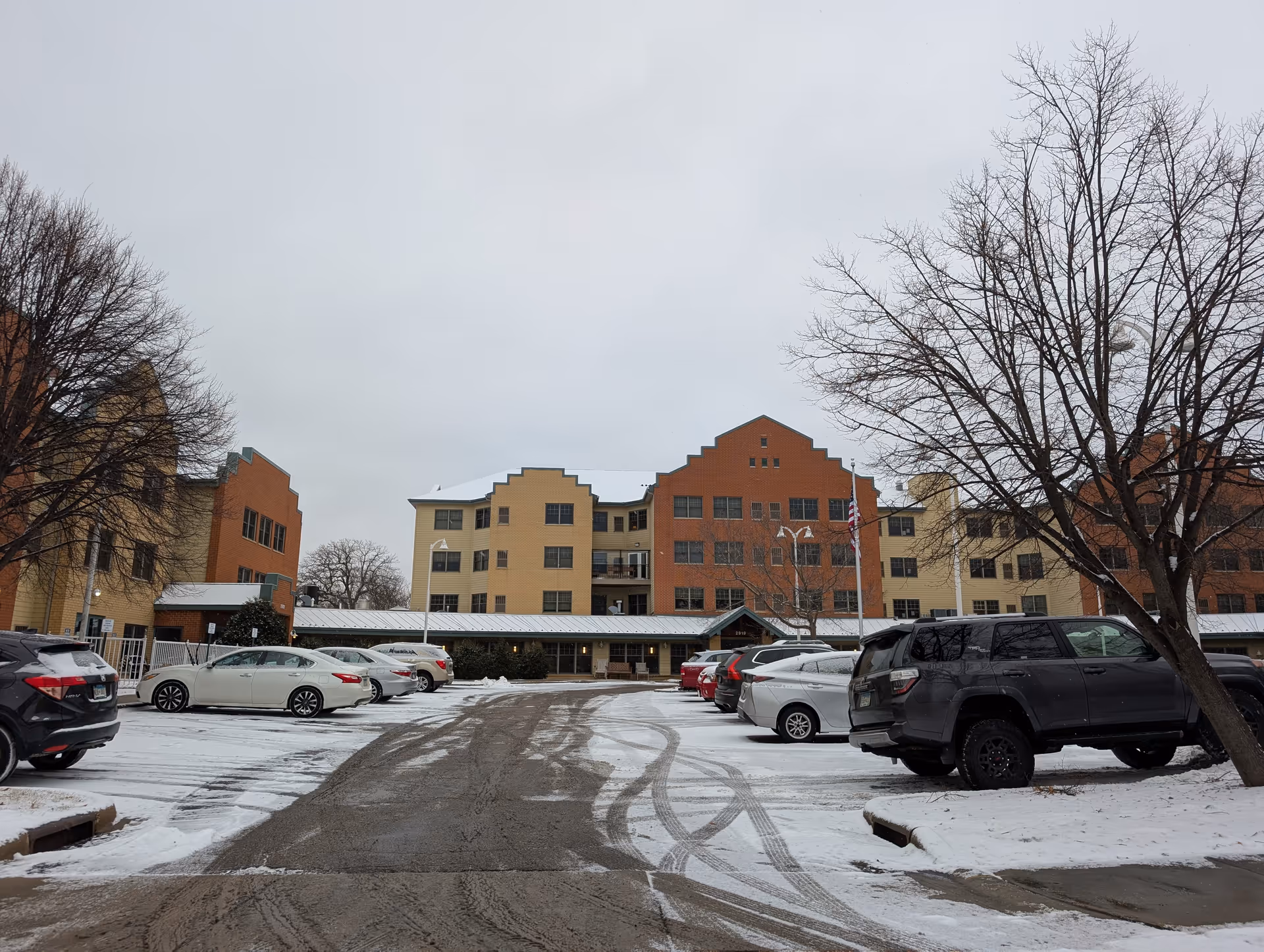 Exterior view of a multi-story senior living facility building with a parking lot in front. Several cars are parked on a snow-covered ground, and leafless trees are visible on both sides of the parking area under an overcast sky.