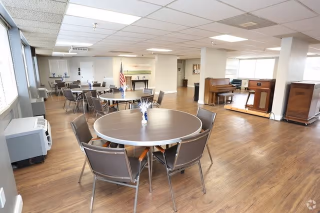 A spacious room with multiple round tables and chairs arranged for dining or social activities. The room has wood flooring, a piano and organ against one wall, large windows letting in natural light, and an American flag near the back wall.