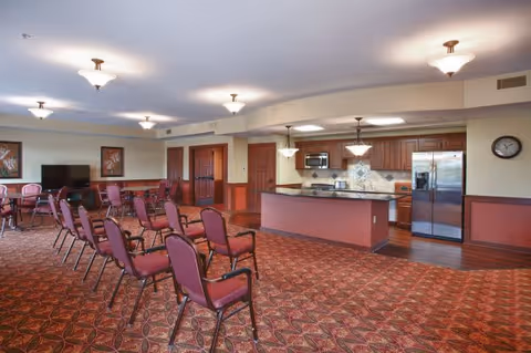 A spacious room with a patterned carpet featuring rows of maroon chairs arranged facing a television on a stand. The room includes a kitchen area with wooden cabinets, stainless steel refrigerator, microwave, and a countertop island. The walls are painted beige with maroon wainscoting, and there are multiple ceiling light fixtures providing illumination.