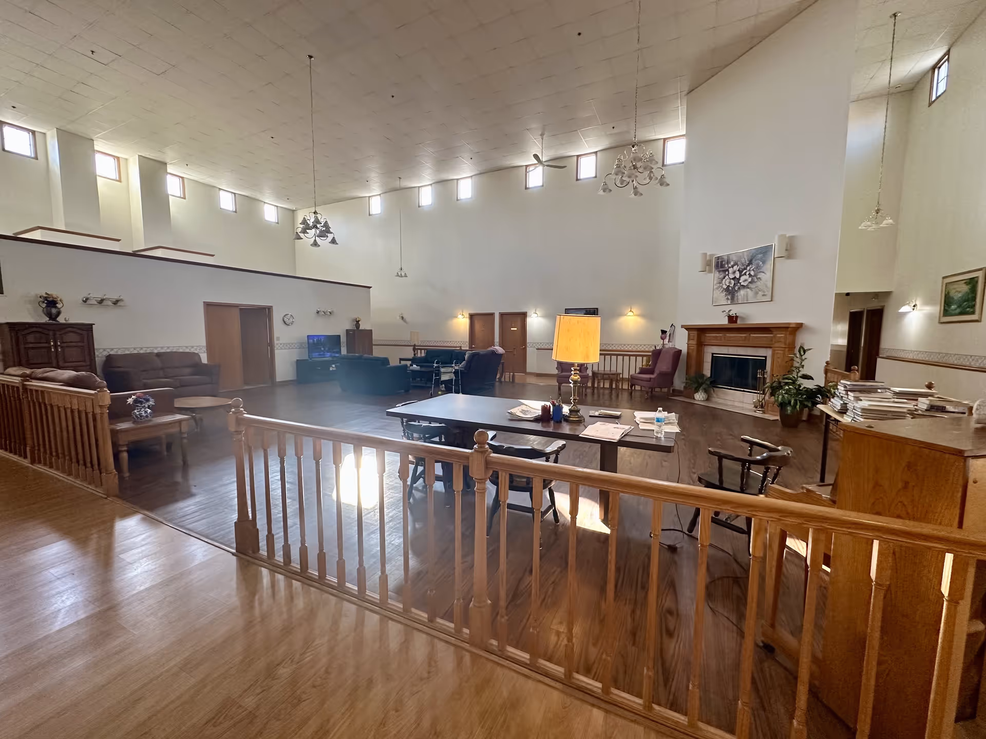 Spacious senior living facility common area with high ceilings and multiple windows near the ceiling allowing natural light. The room features wooden flooring, several seating areas with sofas and chairs, a fireplace with a painting above it, a table with a lamp and papers, and decorative plants. The space is divided by a wooden railing and has a warm, inviting atmosphere.