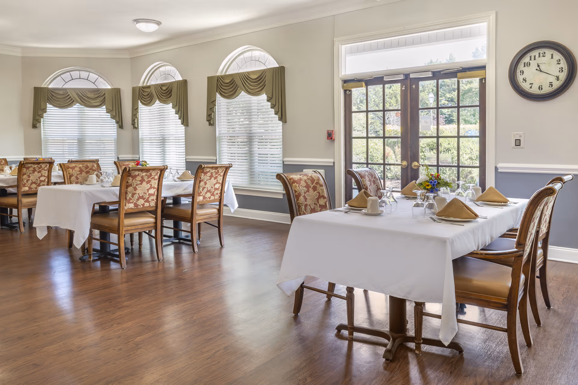 Bright dining room with tables set with white tablecloths, folded napkins, and patterned chairs beside large windows and glass doors.