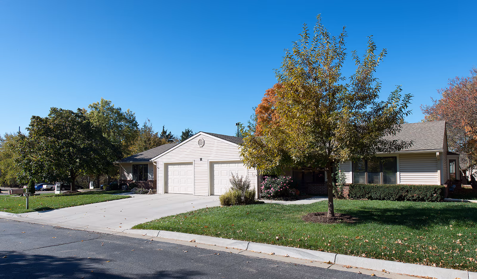 Exterior view of a single-story residential building with beige siding and a two-car garage. The building is surrounded by green grass, trees with autumn foliage, and a clear blue sky overhead.