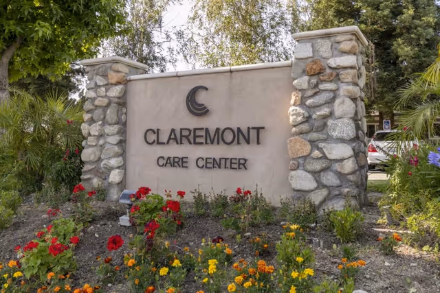 Stone and stucco sign for Claremont Care Center surrounded by colorful flowers and greenery with trees in the background.