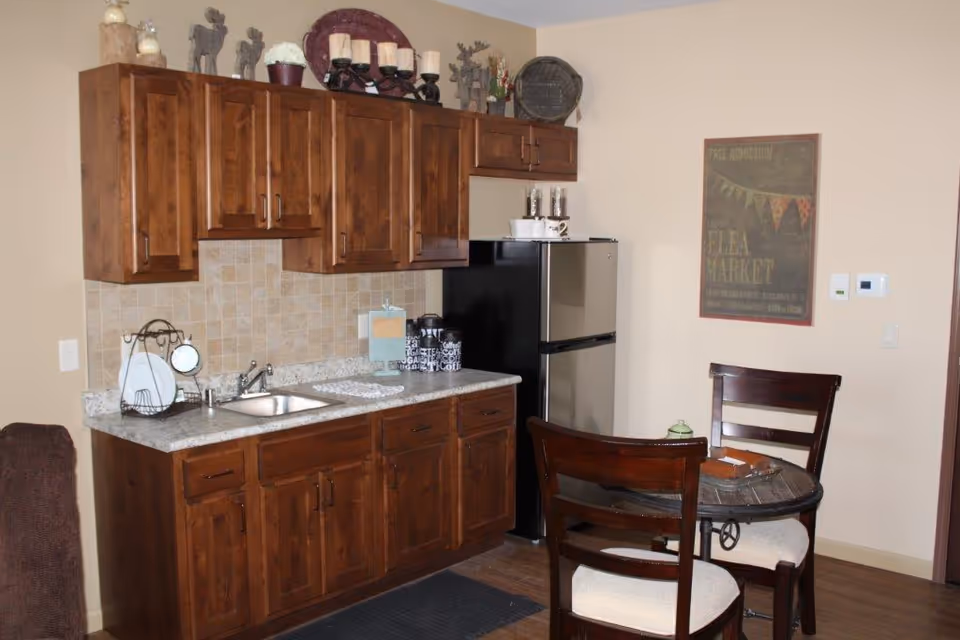 A small kitchen area with wooden cabinets, a granite countertop, a stainless steel sink, and a compact refrigerator. There are decorative items on top of the cabinets and a small round dining table with two wooden chairs in front of the kitchen. A framed poster hangs on the wall next to a thermostat.