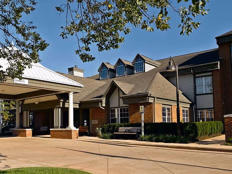 Exterior view of a senior living facility building with a covered entrance supported by white columns, brick walls, multiple windows, and a sloped roof with dormer windows. There are benches, trimmed bushes, a street lamp, and tree branches visible under a clear blue sky.