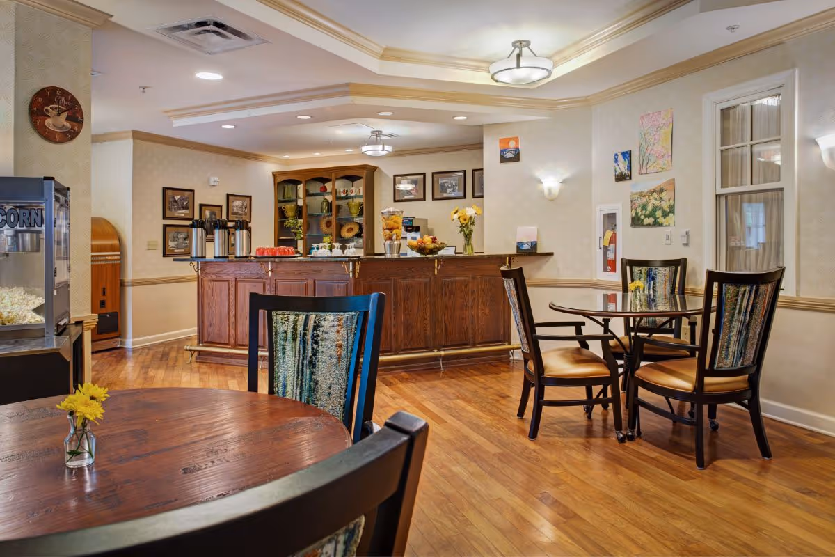 Interior view of a senior living facility dining area with wooden floors, round tables with chairs, a popcorn machine, and a wooden counter with coffee dispensers and snacks. The walls are decorated with framed pictures and paintings, and there are ceiling lights providing warm illumination.